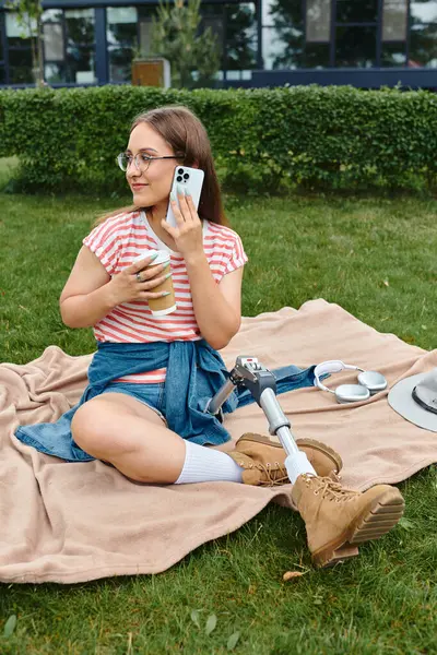 A young woman with a prosthetic leg relaxes on a blanket in a park, enjoying a coffee and a phone call. — Stock Photo