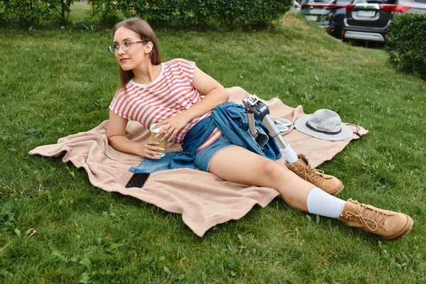 Uma jovem mulher com uma perna protética relaxa em um cobertor em um parque, desfrutando de um café e do sol. — Fotografia de Stock