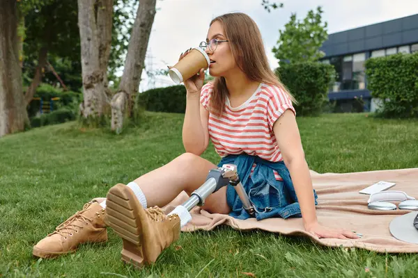 A young woman with a prosthetic leg relaxes on a blanket in a park, enjoying a drink and the warm weather. — Stock Photo