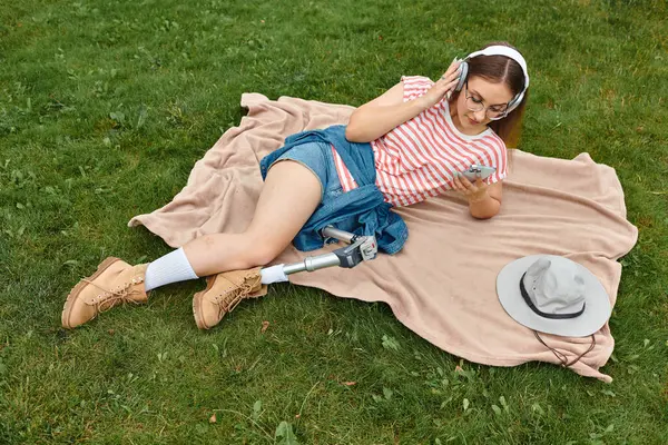 Eine junge Frau im Sommeroutfit entspannt sich auf einer Decke in einem Park, hört Musik über Kopfhörer und scrollt auf ihrem Handy.. — Stockfoto