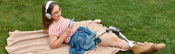 Una joven con una pierna protésica disfruta de un día soleado en el parque, escuchando música y desplazándose en su teléfono. - foto de stock