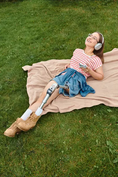 A young woman with a prosthetic leg relaxes on a blanket in a park, listening to music and smiling. — Stock Photo