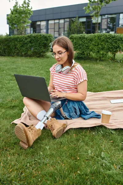 A young woman with a prosthetic leg sits on a blanket in a park, working on her laptop. — Stock Photo
