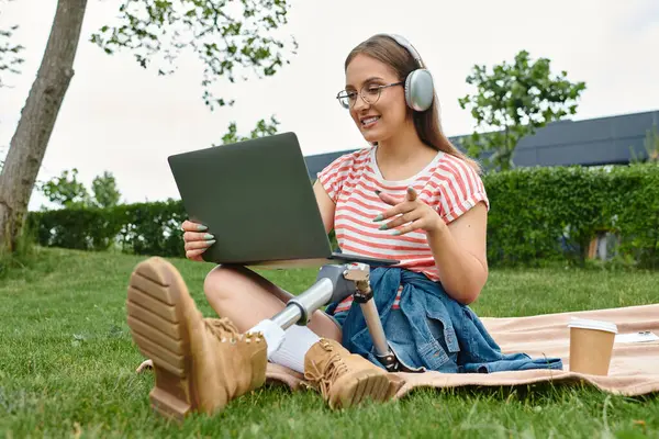 A young woman with a prosthetic leg sits on a blanket in a park, using a laptop and wearing headphones. — Stock Photo