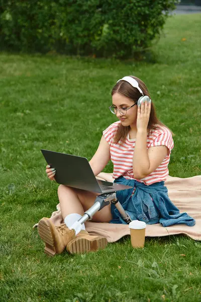 A young woman with a prosthetic leg uses a laptop while sitting in a park, wearing headphones and a summery outfit. — Stock Photo