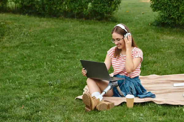 Uma jovem mulher com uma perna protética senta-se em um cobertor em um parque, trabalhando em seu laptop e ouvindo música com fones de ouvido. — Fotografia de Stock
