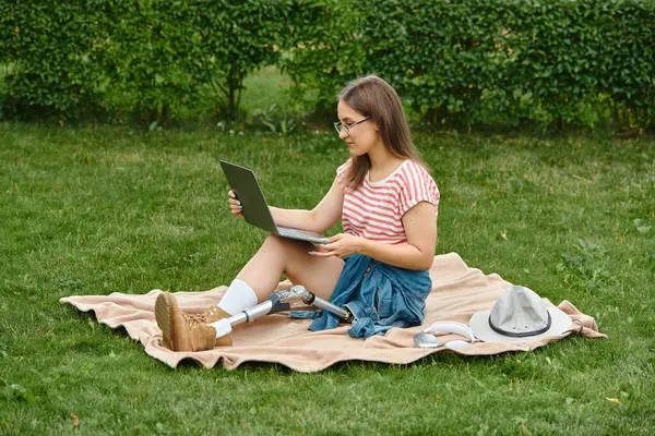 A young woman with a prosthetic leg uses her laptop while sitting on a blanket in a park. — Stock Photo