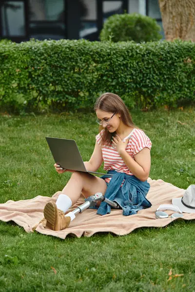 A young woman with a prosthetic leg sits on a blanket in a park, smiling as she uses her laptop. — Stock Photo