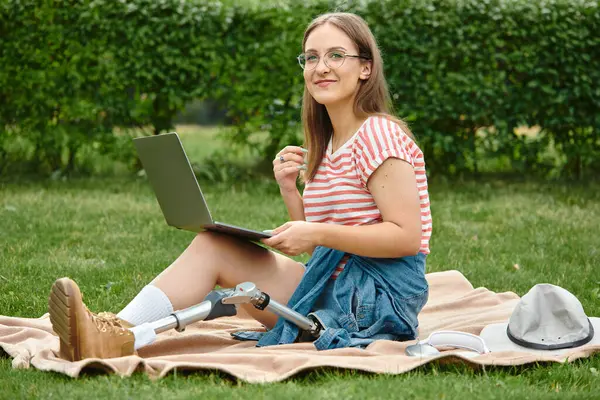 Uma jovem mulher com uma perna protética senta-se em um cobertor em um parque, usando um laptop. — Fotografia de Stock