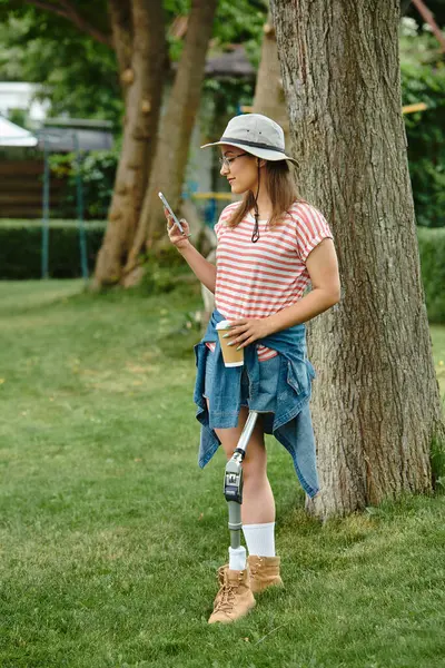 Eine junge Frau mit Beinprothese genießt einen sonnigen Tag im Park und kontrolliert ihr Handy, während sie eine Tasse Kaffee in der Hand hält. — Stockfoto