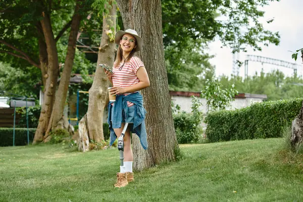 Giovane donna in abito estivo sorridente con fiducia, appoggiata ad un albero in un parco con una tazza di caffè — Foto stock