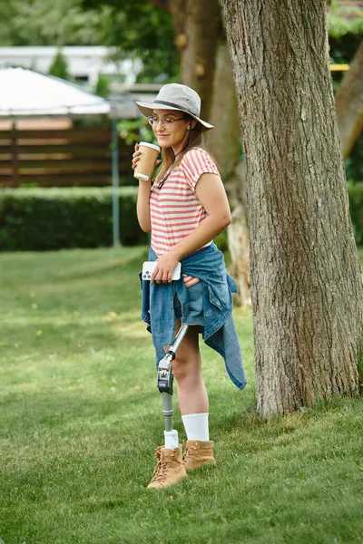 Uma jovem mulher com uma perna protética fica perto de uma árvore, desfrutando de uma xícara de café. — Fotografia de Stock