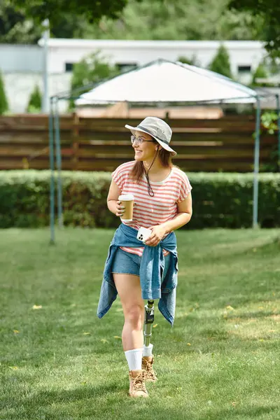 A young woman with a prosthetic leg walks through a park, enjoying a sunny day with a cup of coffee in hand. — Stock Photo