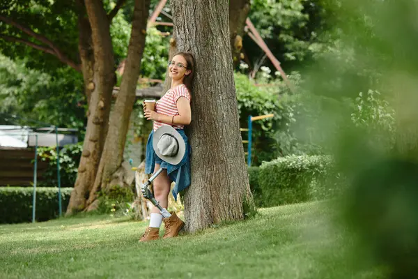 A young woman with a prosthetic leg leans against a tree, enjoying a coffee in the park. — Stock Photo