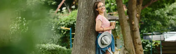 A young woman in a summer outfit leans against a tree in a park, enjoying a cup of coffee. — Stock Photo