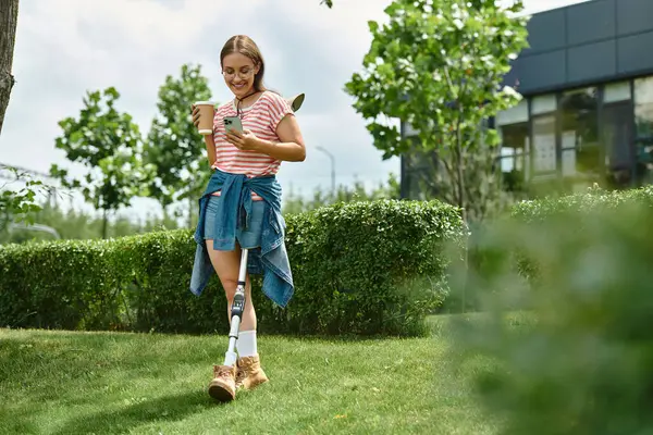A young woman with a prosthetic leg smiles while looking at her phone and holding a cup of coffee in a summer park. — Stock Photo
