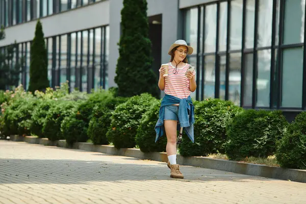 Uma jovem mulher com uma perna protética caminha em um caminho de tijolo através de um parque da cidade, segurando uma xícara de café e um smartphone em suas mãos. — Fotografia de Stock