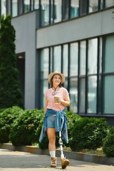 A young woman with a prosthetic leg walks through a city park, holding a cup of coffee and smiling. — Stock Photo