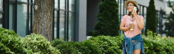 Una giovane donna cammina attraverso un parco, godendo la giornata estiva. E 'al telefono e beve qualcosa.. — Foto stock