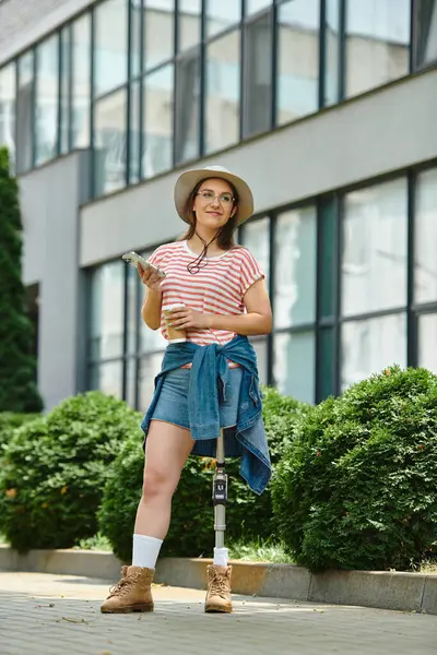 A young woman with a prosthetic leg enjoys a sunny afternoon in the city, wearing a stylish summer outfit and carrying a cup of coffee. — Stock Photo