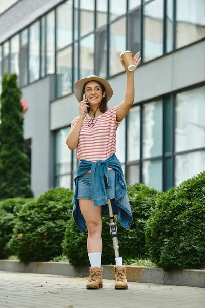 Una joven con una pierna protésica camina en un parque de la ciudad, sosteniendo una taza de café en una mano y hablando por teléfono en la otra. - foto de stock