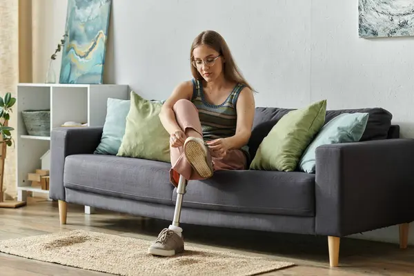 A young woman with a prosthetic leg sits on a couch in her living room, tying her shoe. — Stock Photo