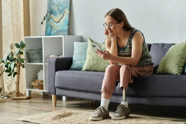 A young woman with a prosthetic leg relaxes on a couch in her living room, looking at her phone. — Stock Photo