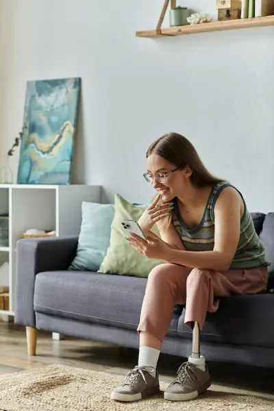 A young woman with a prosthetic leg smiles while looking at her phone while sitting on a couch in her living room. — Stock Photo