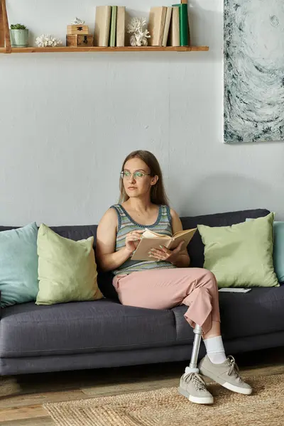 A young woman with a prosthetic leg sits on a sofa in her living room, reading a book and gazing thoughtfully out of the window. — Stock Photo