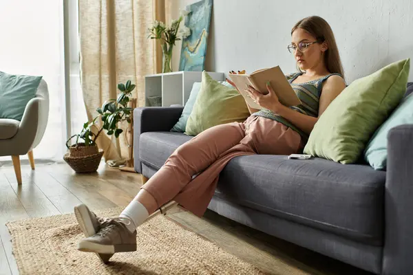 A young woman with a prosthetic leg relaxes on a couch in her living room, engrossed in a book. — Stock Photo