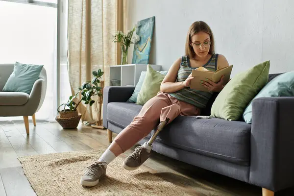 A young woman with a prosthetic leg relaxes on a gray sofa, engrossed in a book. — Stock Photo