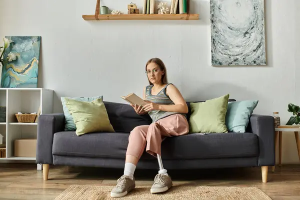 Una mujer joven se sienta en un sofá gris en su sala de estar, sosteniendo un libro y mirando pensativamente a la distancia. — Stock Photo