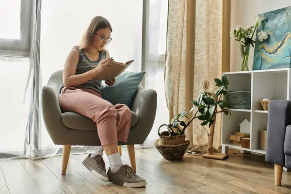 A young woman with a prosthetic leg sits in a comfortable armchair in a modern living room, reading a book. — Stock Photo