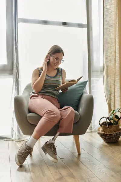 A young woman with a prosthetic leg sits in a comfortable armchair, reading a book while wearing glasses. — Stock Photo