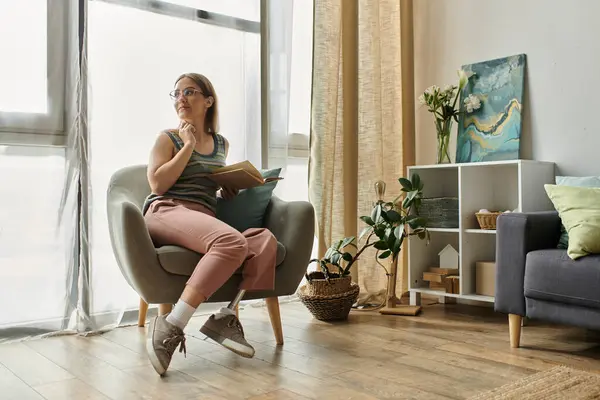 Eine junge Frau mit Beinprothese sitzt in einem bequemen Sessel in ihrem Wohnzimmer, verloren in Gedanken. — Stockfoto