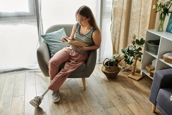 Eine junge Frau mit Beinprothese sitzt in einem bequemen Sessel und liest in ihrem modernen Wohnzimmer ein Buch. — Stockfoto