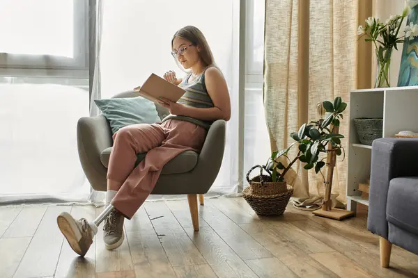 A young woman with a prosthetic leg relaxes in a comfortable chair, engrossed in a book. Natural light floods the modern living room. — Stock Photo