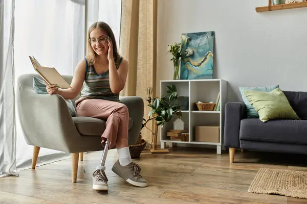 A young woman with a prosthetic leg sits in a comfortable chair and reads a book in her modern living room. — Stock Photo