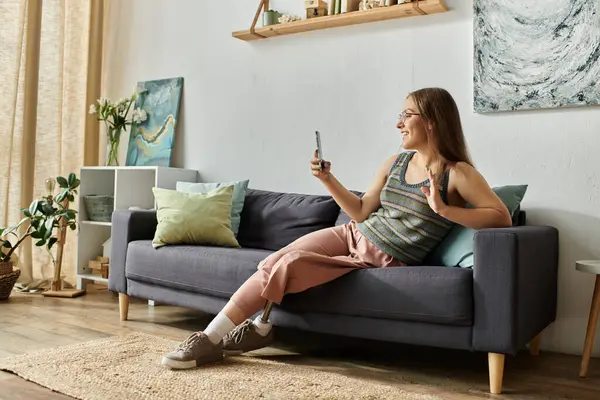 A young woman with a prosthetic leg sits on a couch in her modern living room, smiling and waving as she uses her phone. — Stock Photo