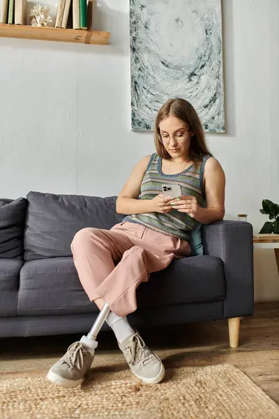 A young woman with a prosthetic leg relaxes on a couch in her modern living room, engrossed in her phone. — Stock Photo