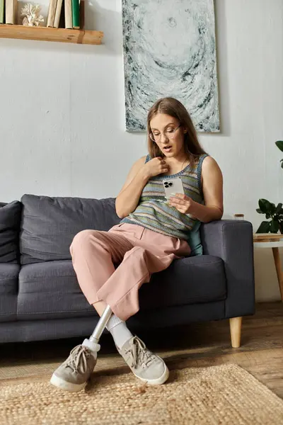 A young woman with a prosthetic leg sits on a couch in her living room, surprised by something she is reading on her phone. — Stock Photo