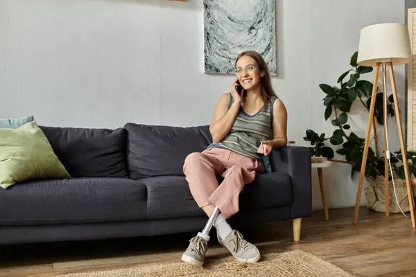 Una giovane donna si siede su un divano nel suo salotto moderno, chiacchierando sul suo telefono. Sorride mentre si gode il suo tempo a casa. — Foto stock