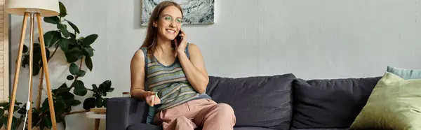 A young woman sits on a couch in her living room, talking on her phone and smiling. — Stock Photo