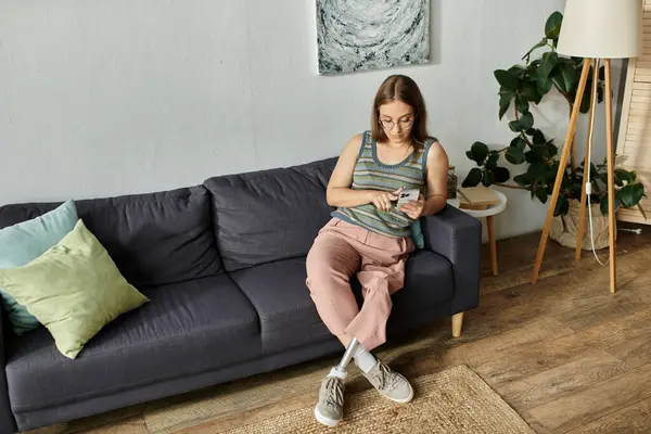 A young woman with a prosthetic leg sits on a sofa in her living room, looking at her phone. — Stock Photo