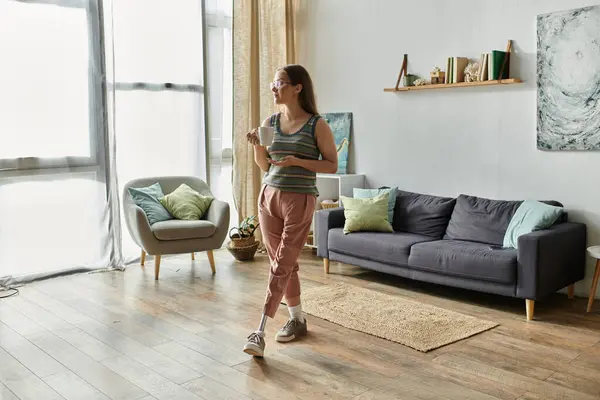 A young woman stands in her modern living room, holding a mug of coffee and looking out a large window. — Stock Photo