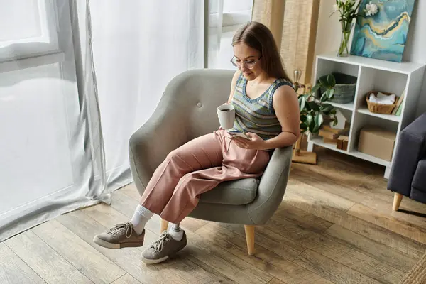 Eine junge Frau mit Beinprothese sitzt in einem bequemen Sessel, hält eine Tasse Kaffee in der Hand und blättert auf ihrem Handy.. — Stockfoto