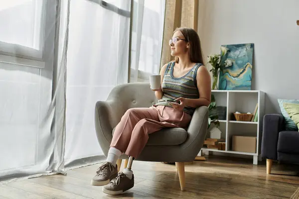 Eine junge Frau mit Beinprothese sitzt in einem bequemen Stuhl in ihrem Wohnzimmer, hält eine Tasse in der Hand und schaut aus dem Fenster. — Stockfoto