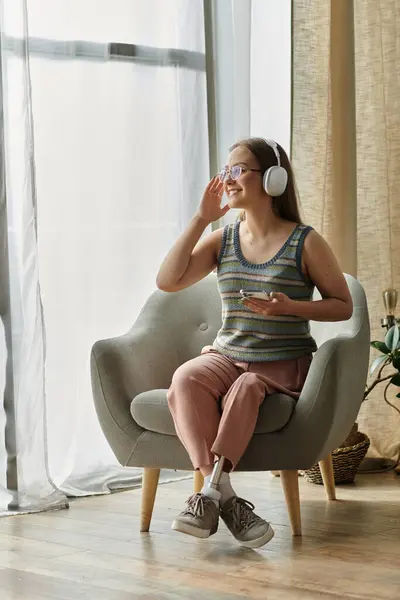 A young woman with a prosthetic leg sits in a modern living room, listening to music with headphones on. — Stock Photo