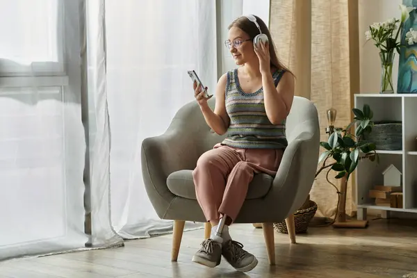 Una joven con una pierna protésica se relaja en su moderna sala de estar, escuchando música y desplazándose por su teléfono. - foto de stock