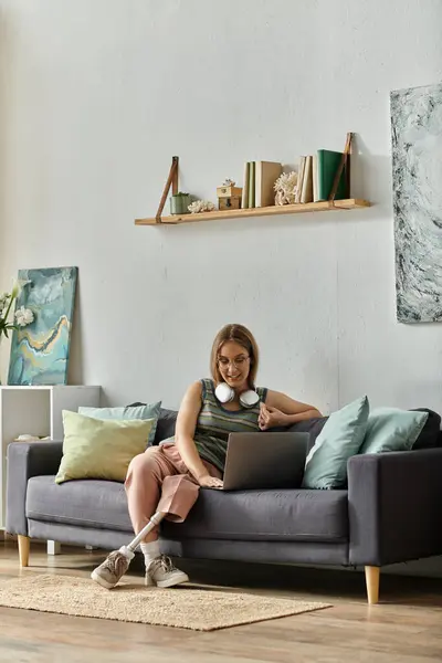 A young woman with a prosthetic leg sits comfortably on her couch, using a laptop. — Stock Photo
